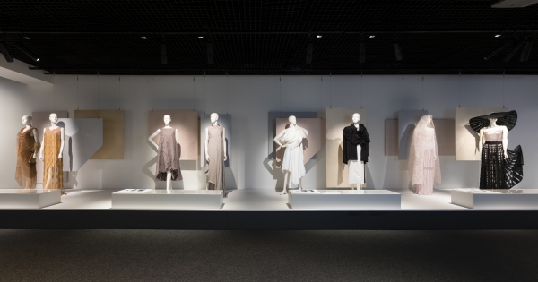 Eight female mannequins dressed in garments designed and made by the four students profiled in Student Fashion 2019. The mannequins are grouped in pairs, and stand in a straight line atop a long rectangular exhibition plinth. Four low rectangular showcases are positioned in front of the mannequins, containing creative process material.
